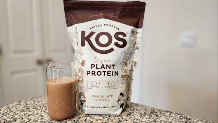 A bag of KOS plant protein in Chocolate flavor and a protein shake in a glass cup on a granite counter